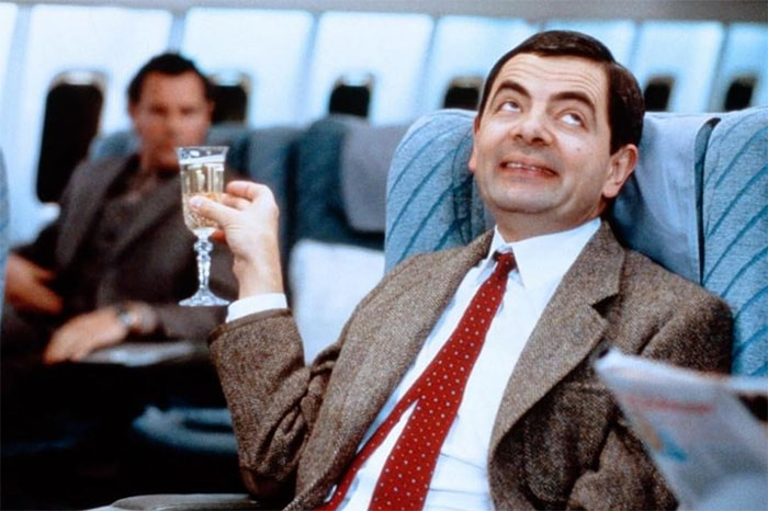 Actor embodying a classic role, holding a drink in an airplane seat.