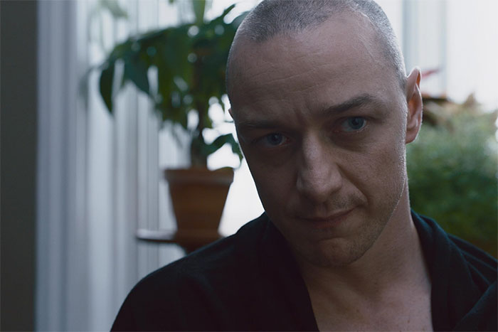 Actor delivering a flawless performance with a serious expression, standing near a window with plants in the background.