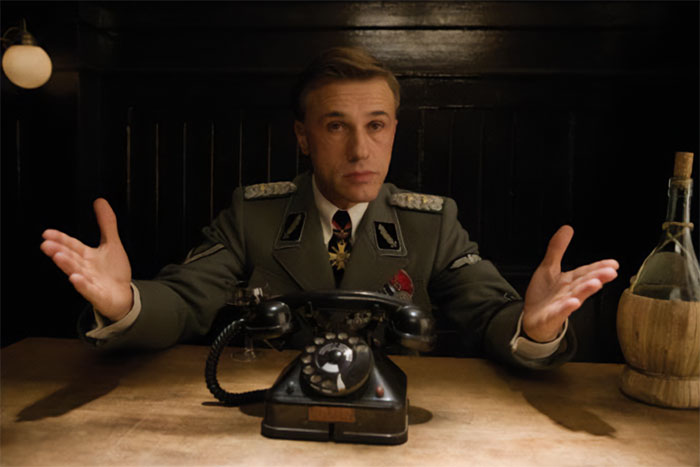 Actor in a military uniform sits at a desk with an old-fashioned phone, portraying a flawless role.