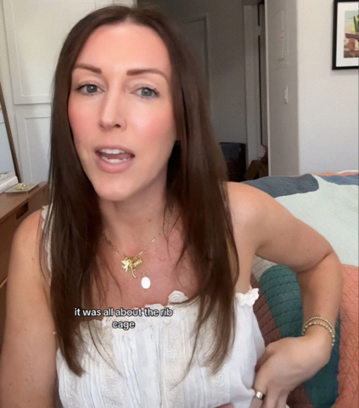 Woman discussing tattoo trends, sitting casually indoors, wearing a white top and gold necklace.