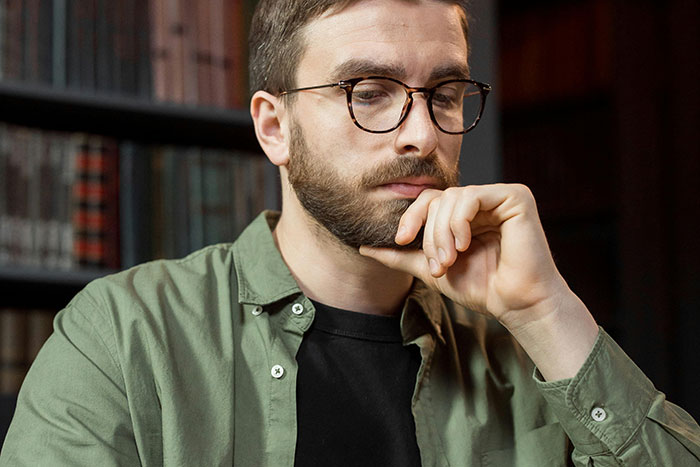 Man thinking deeply in a library, wearing glasses and a green shirt, reflecting on essential life skills.