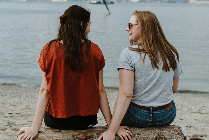 Two women sitting on a beach log, discussing essential life skills and enjoying the view.