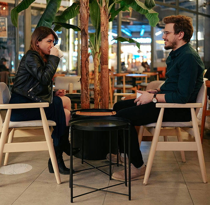 Woman on a date in a cafe, sipping coffee, with a man talking, surrounded by plants in a cozy setting.