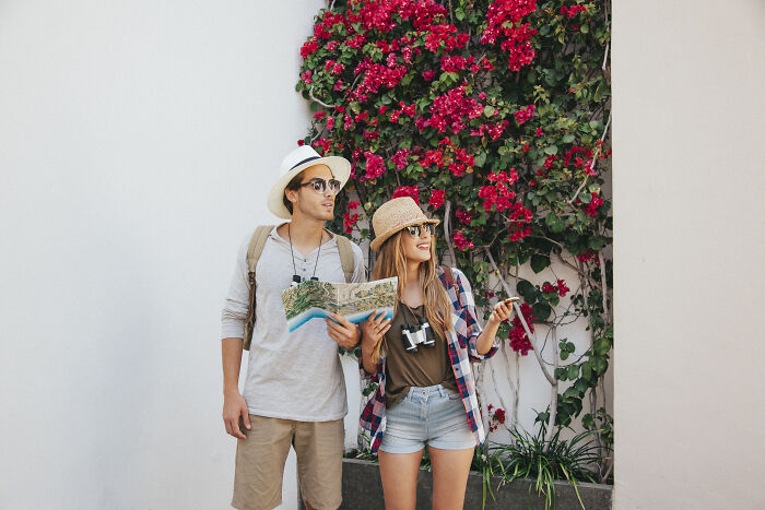 Two tourists with a map and phone, pondering directions, standing in front of a vibrant wall of red flowers.