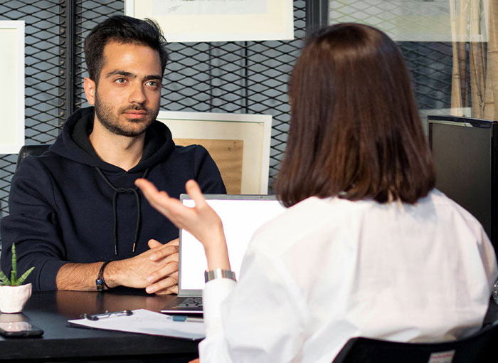 Two people discussing Hollywood secrets in an office, with one wearing a black hoodie.