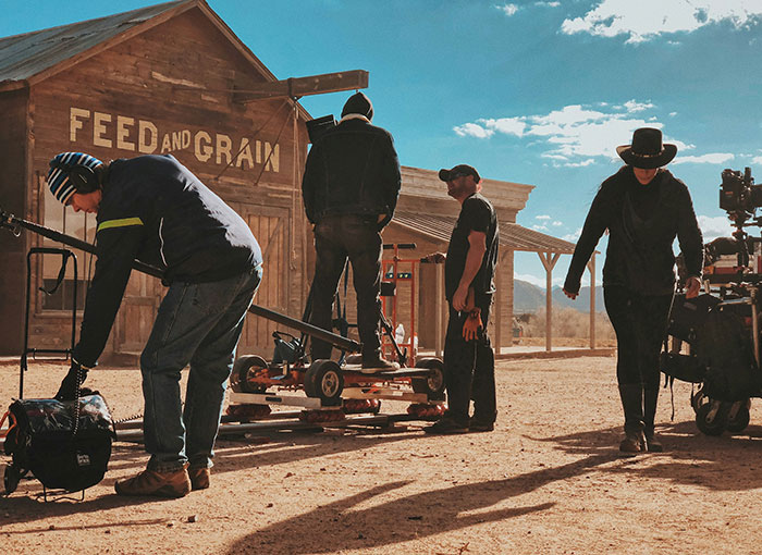 Crew on a Hollywood set in desert setting, adjusting film equipment in front of rustic building labeled "Feed and Grain."