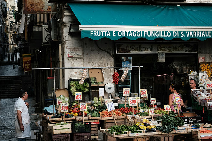 Fruit and vegetable market in a busy European street, with vendors and colorful produce displays.