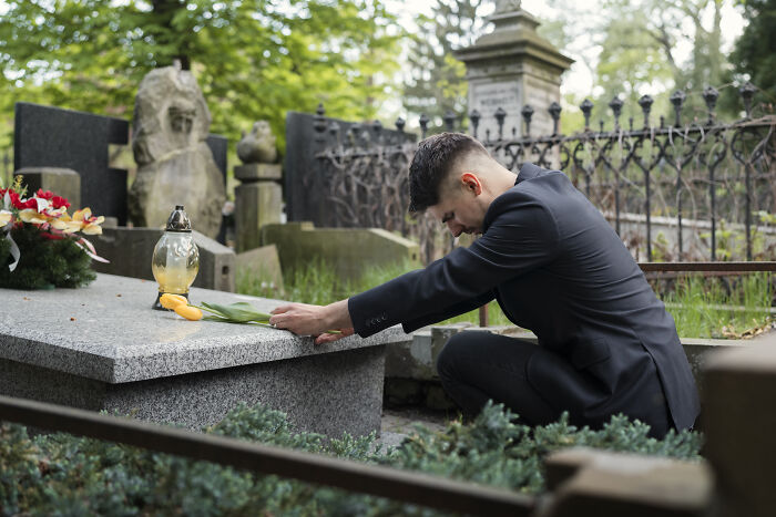 Man in black suit kneeling at a grave, placing flowers during a solemn moment at a cemetery.