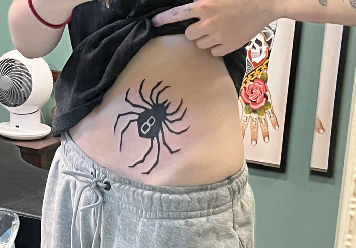 Woman showing a large spider tattoo on her side, dressed in gray sweatpants, with artwork in the background.