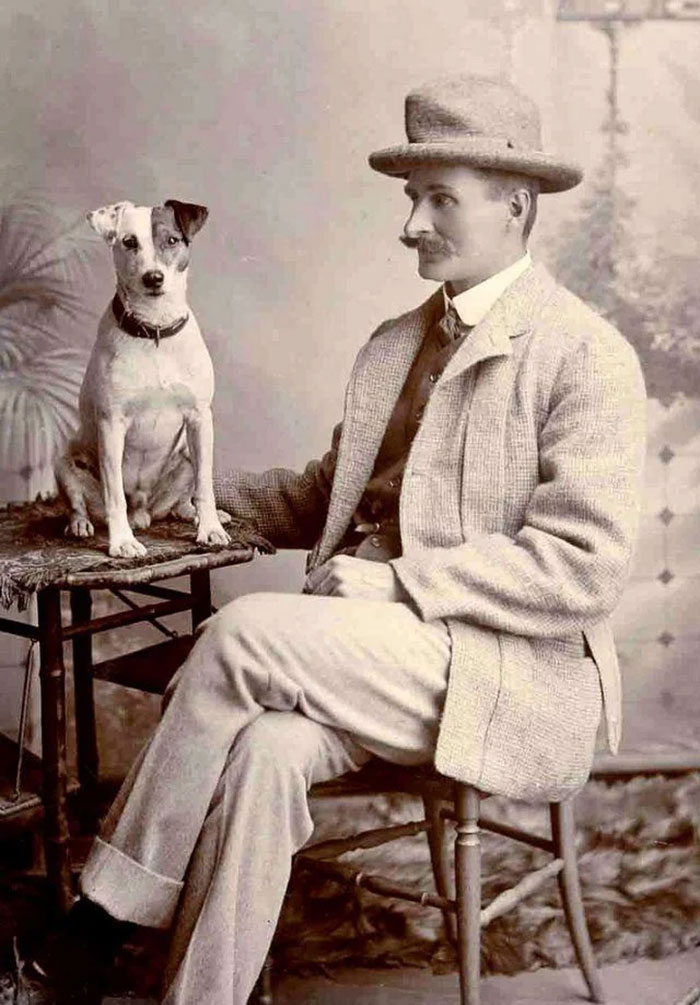 Vintage photo of a man in a suit and hat sitting with a dog on a small table, capturing family and pet bond.