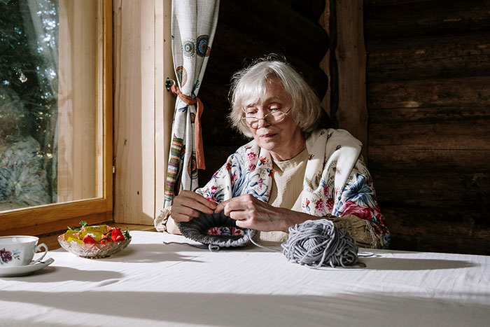 Senior woman knitting alone by a sunny window, with a cup of tea and fruit on the table.
