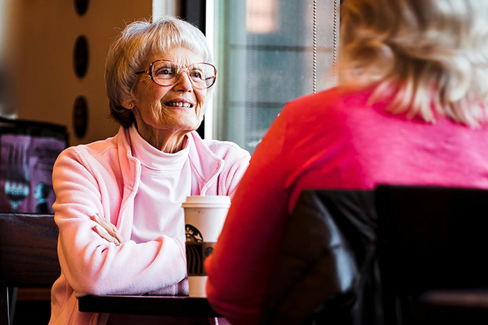 Senior woman smiling and chatting over coffee at a cafe, wearing glasses and a pink jacket.