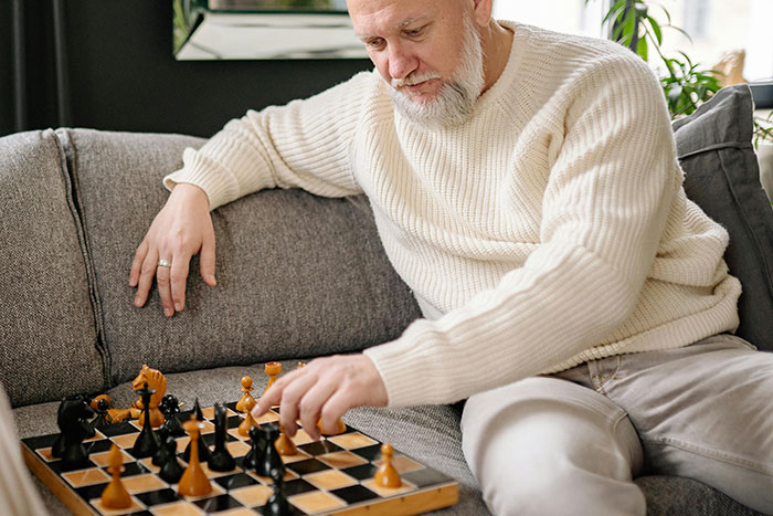 Senior man in a cozy sweater playing chess alone on a gray sofa.