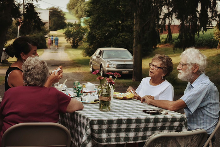 Seniors enjoying a conversation at an outdoor table, surrounded by greenery and parked cars.