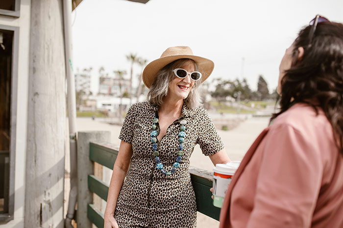 Senior woman in a hat and sunglasses smiling, talking to a friend by the beach.