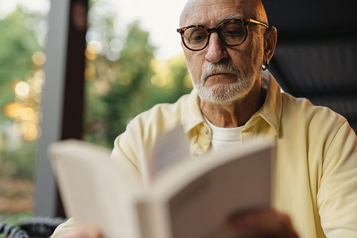 Senior man with glasses reading a book outdoors, wearing a yellow shirt, immersed in thought.