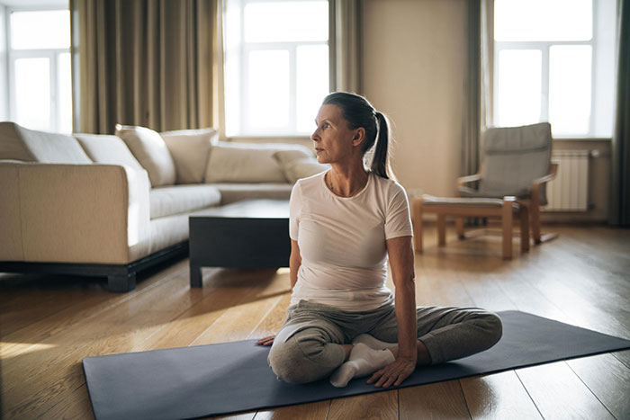 Senior woman doing yoga alone in a living room, seated on a mat in a sunlit space.