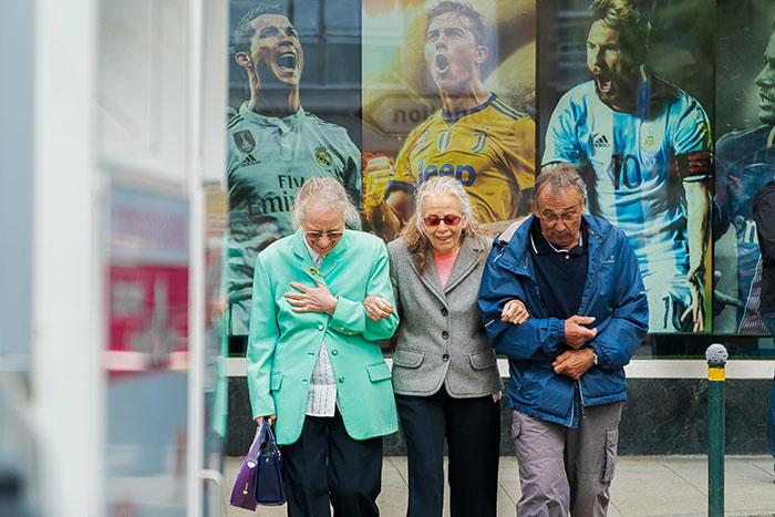 Three seniors walk together, arms linked, in front of a sports-themed mural, highlighting connection and companionship.