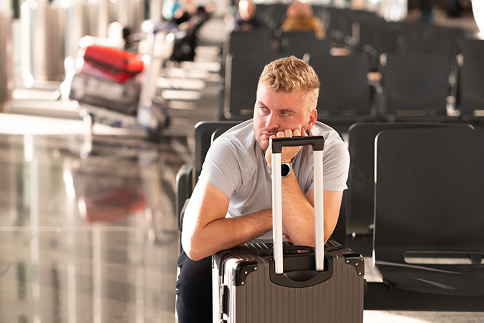 Man at airport lounges on suitcase, appearing frustrated, amid empty seats.