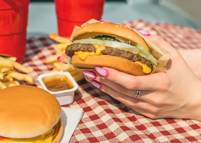 A person with pink nails holding a cheeseburger over a checkered tablecloth, with fries and a drink in the background.