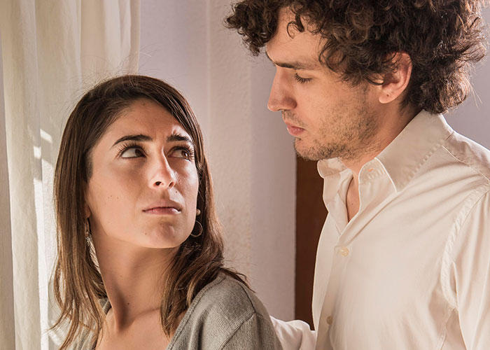 A man and woman discussing harsh life realities indoors, both with serious expressions.