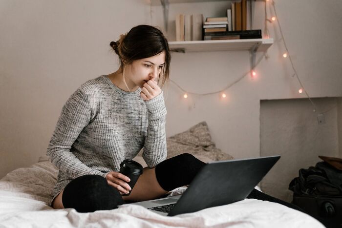 Woman in cozy sweater sitting on bed, holding coffee, engaged with a laptop, fairy lights in background.