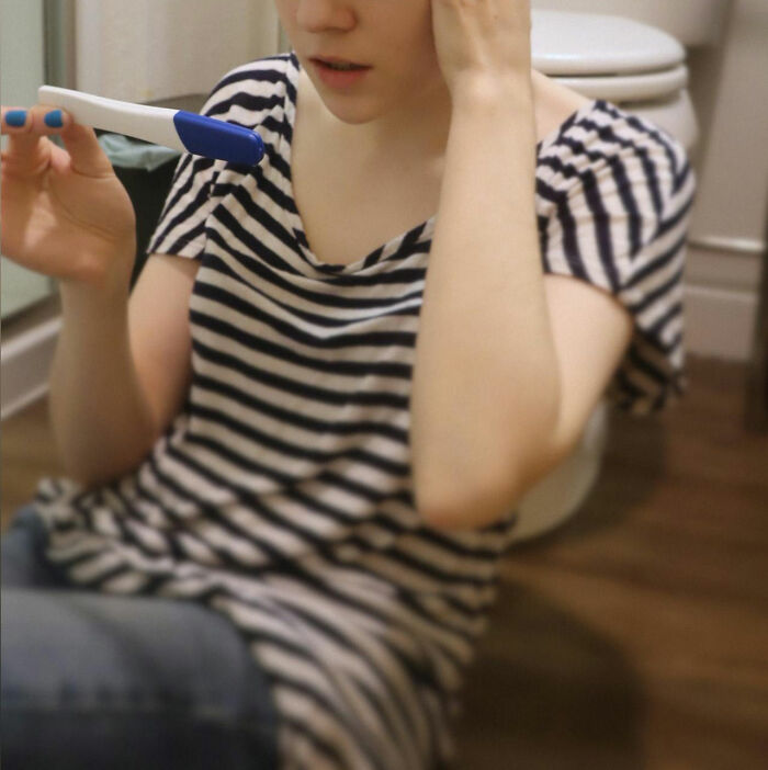 A woman in a striped shirt sitting on a bathroom floor, looking at a pregnancy test; concept of family secrets.