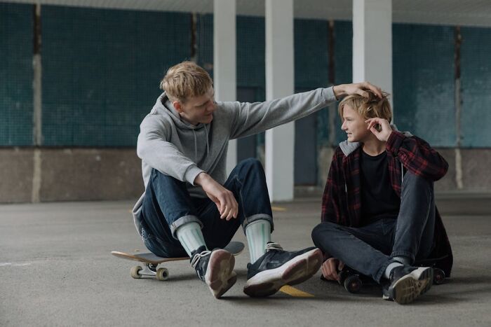 Two brothers sitting on skateboards in a parking garage, sharing a light moment, hinting at family secrets.