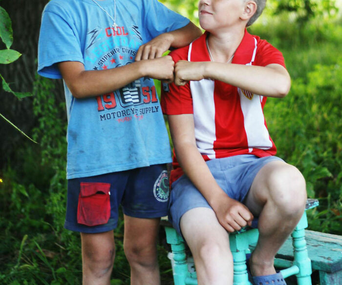 Two boys in casual outfits are sitting and fist-bumping in a garden, symbolizing friendship and family secrets.