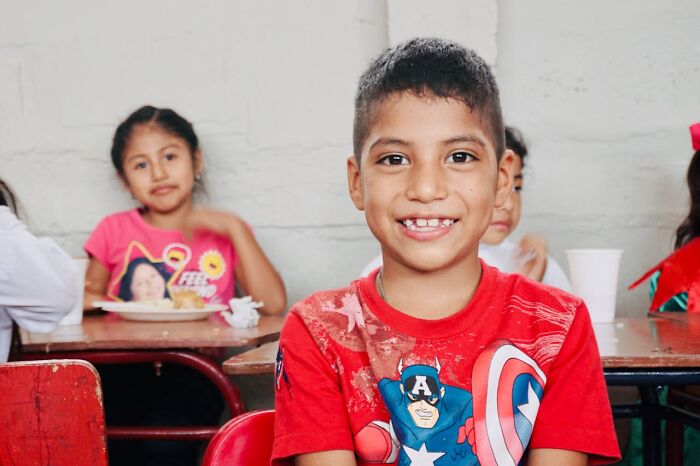 Child smiling in a classroom, wearing a superhero t-shirt, with classmates in the background.