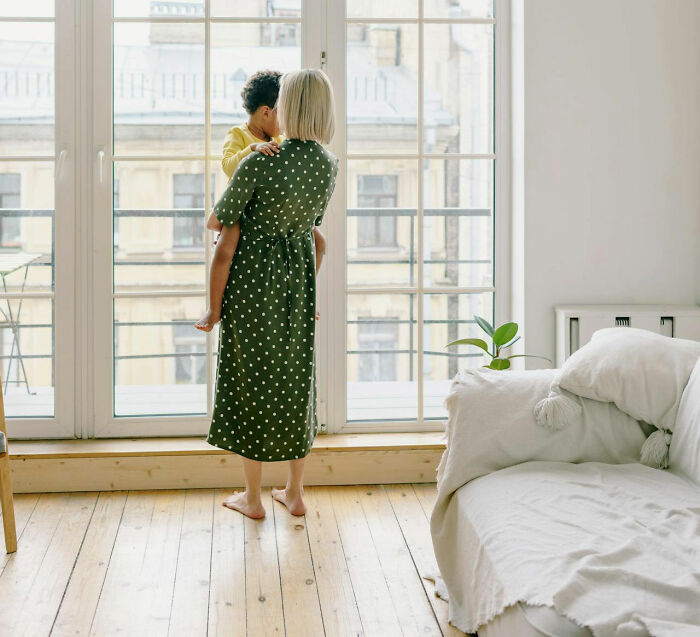 Woman holding a child by a large window, symbolizing family secrets and bonding moments.