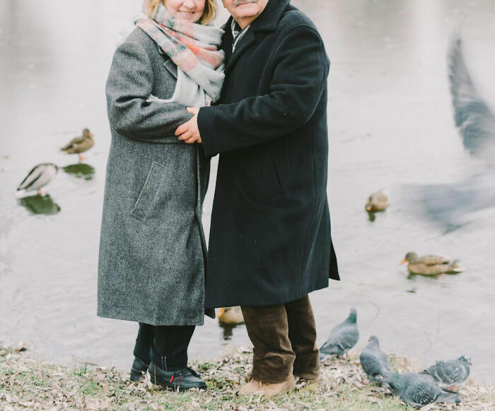 Elderly couple embracing by a pond, wearing winter coats, surrounded by ducks and pigeons, symbolizing family secrets.