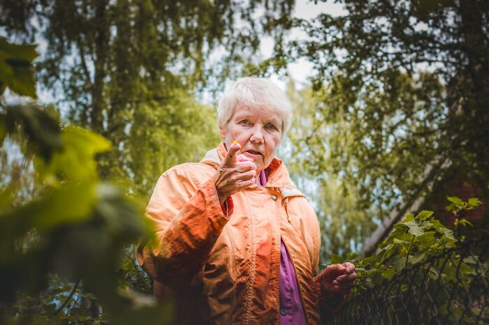 Elderly woman in orange jacket outdoors, surrounded by trees, gesturing with hand, evoking family-secrets theme.