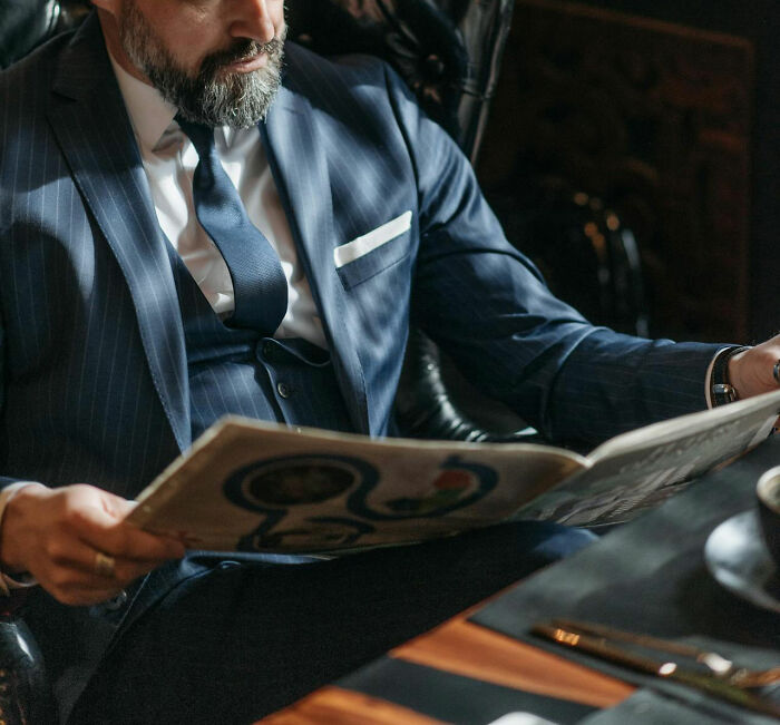Man in a suit reading a newspaper, uncovering family-secrets, in a cozy, dimly-lit office setting.