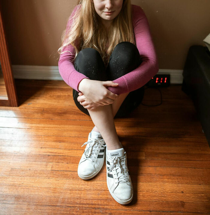 Person in a pink sweater sitting on a wooden floor near a clock, reflecting on family secrets.