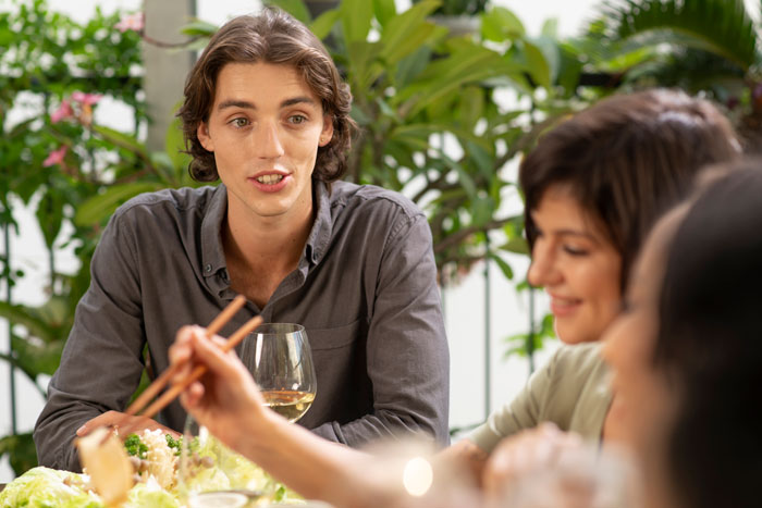 Teen sitting at a dinner table, looking away while others are engaged in conversation, surrounded by greenery.