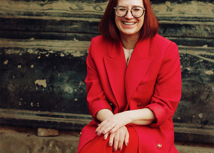 Woman in bright red suit sitting, smiling in front of a textured wall. Woman in bright red suit sitting, smiling in front of a textured wall.