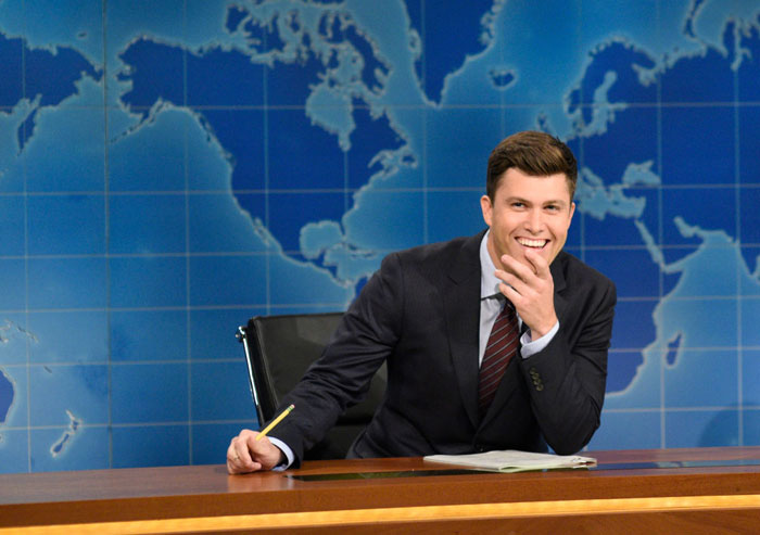 Colin Jost, Scarlett Johansson&rsquo;s husband, sitting at a news desk, smiling confidently, wearing a suit and tie with a map backdrop.