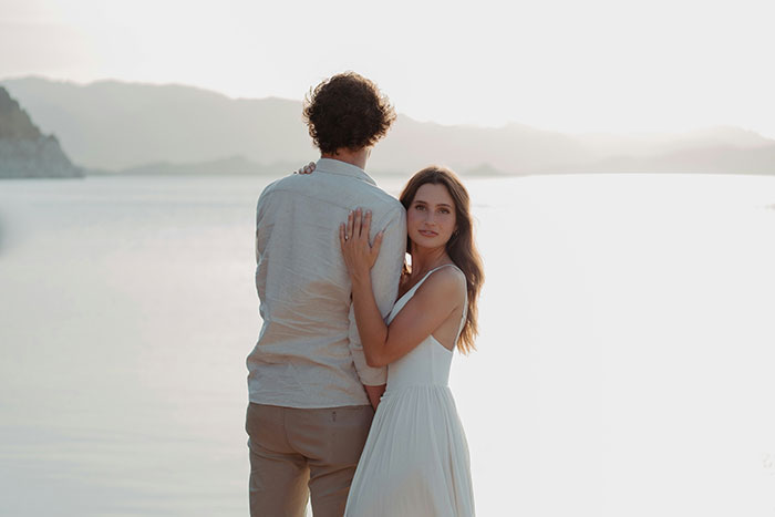 Couple standing by the water at sunset, woman in white dress looking at the camera, illustrating marriage themes.