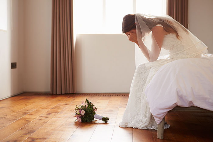 Bride in distress sitting on a bed with her wedding bouquet on the floor, reflecting "wedding tax" chaos.