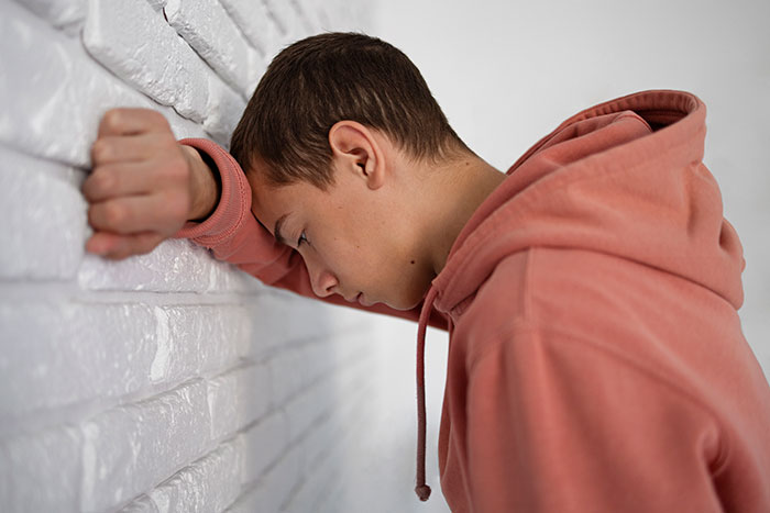 Teen in a peach hoodie leans forehead against a white brick wall, appearing deep in thought.