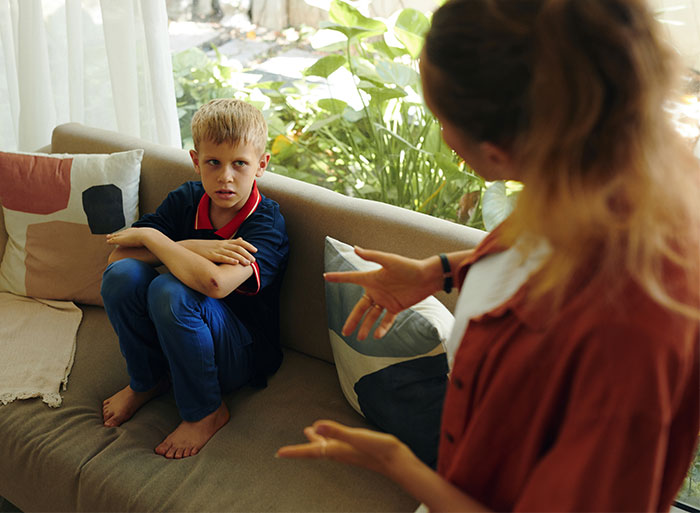 A boy sits on a couch looking upset, while a woman stands nearby gesturing with her hands, relating to babysitter challenges.