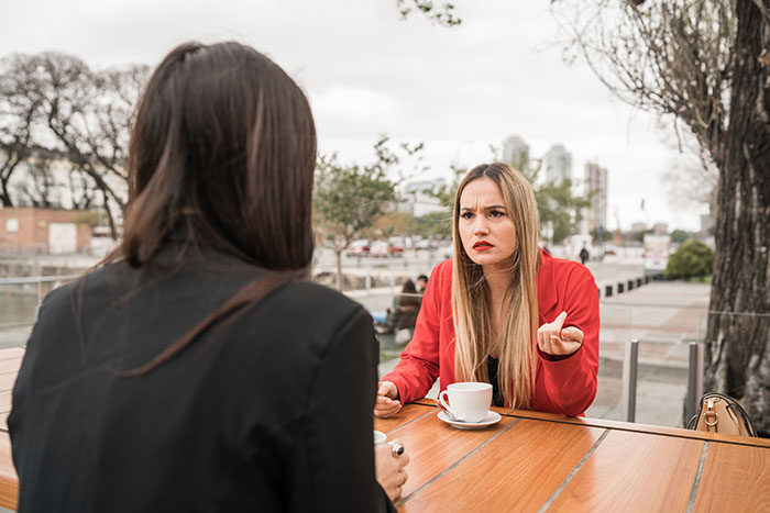 Two women having a tense conversation outdoors, one looking frustrated over a cup of coffee. Two women having a tense conversation outdoors, one looking frustrated over a cup of coffee.