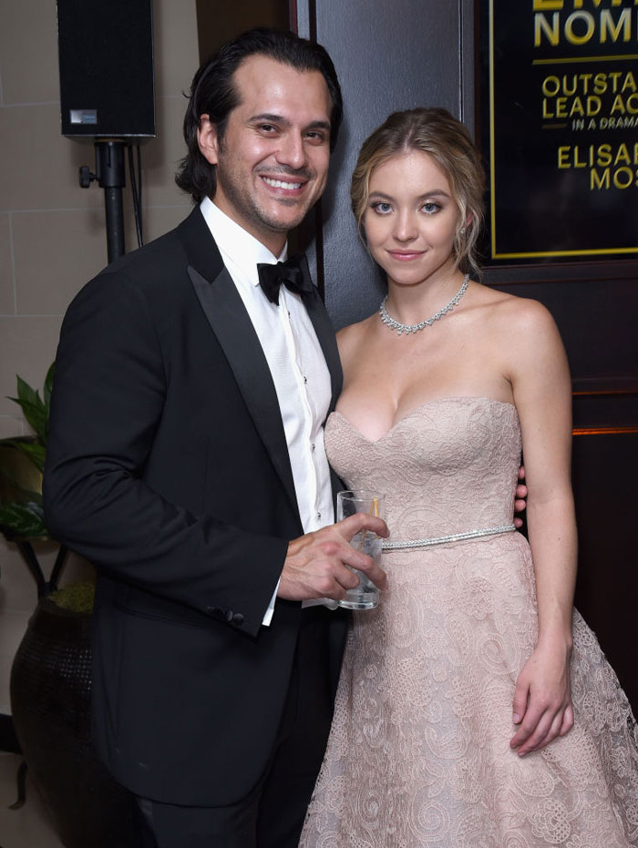 Sydney Sweeney, wearing a strapless light pink lace gown with a delicate necklace, poses beside Jonathan Davino, dressed in a black tuxedo and bow tie. They smile together at an event. Sydney Sweeney, wearing a strapless light pink lace gown with a delicate necklace, poses beside Jonathan Davino, dressed in a black tuxedo and bow tie. They smile together at an event.