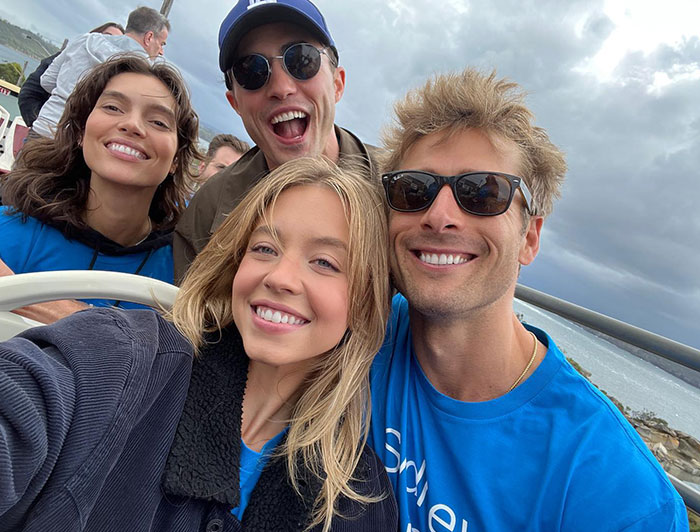 Sydney Sweeney poses with friends outdoors, all smiling and looking cheerful. Sydney is dressed in a dark jacket over a blue shirt, while her friends wear blue shirts and sunglasses, with one in a cap. They appear to be enjoying a casual day together with an ocean view in the background, under a cloudy sky. Sydney Sweeney poses with friends outdoors, all smiling and looking cheerful. Sydney is dressed in a dark jacket over a blue shirt, while her friends wear blue shirts and sunglasses, with one in a cap. They appear to be enjoying a casual day together with an ocean view in the background, under a cloudy sky.