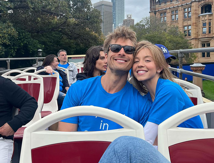 Sydney Sweeney and a friend sit together on an open-top tour bus, both wearing matching blue shirts. Sydney leans affectionately on her friend, smiling broadly, while he wears sunglasses and grins. The setting appears to be in a city, with skyscrapers and historic buildings visible in the background. Other passengers can be seen enjoying the scenic ride. Sydney Sweeney and a friend sit together on an open-top tour bus, both wearing matching blue shirts. Sydney leans affectionately on her friend, smiling broadly, while he wears sunglasses and grins. The setting appears to be in a city, with skyscrapers and historic buildings visible in the background. Other passengers can be seen enjoying the scenic ride.