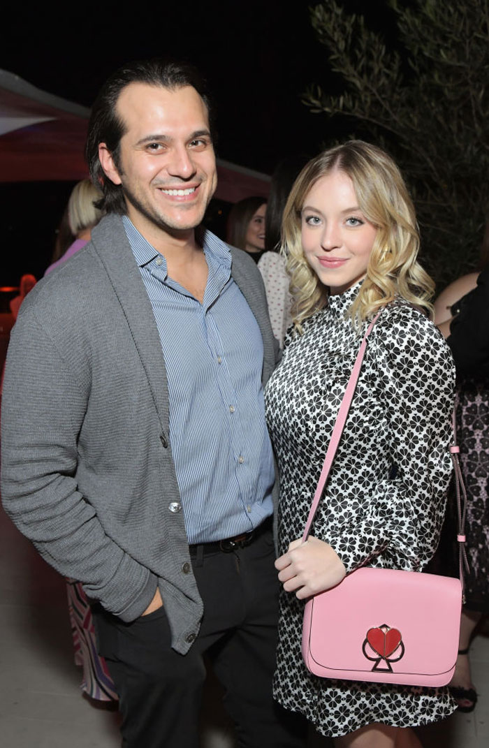 Sydney Sweeney, dressed in a black-and-white patterned long-sleeve dress and carrying a pink bag with a heart design, stands beside Jonathan Davino, who is wearing a blue striped shirt with a gray cardigan. They smile together at an event. Sydney Sweeney, dressed in a black-and-white patterned long-sleeve dress and carrying a pink bag with a heart design, stands beside Jonathan Davino, who is wearing a blue striped shirt with a gray cardigan. They smile together at an event.