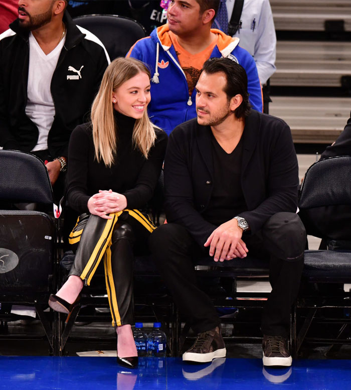 Sydney Sweeney, dressed in a black turtleneck and black pants with yellow side stripes, sits courtside at a sports event, smiling at Jonathan Davino, who is dressed in black attire with a watch on his wrist. They appear engaged in conversation amidst the crowd. Sydney Sweeney, dressed in a black turtleneck and black pants with yellow side stripes, sits courtside at a sports event, smiling at Jonathan Davino, who is dressed in black attire with a watch on his wrist. They appear engaged in conversation amidst the crowd.