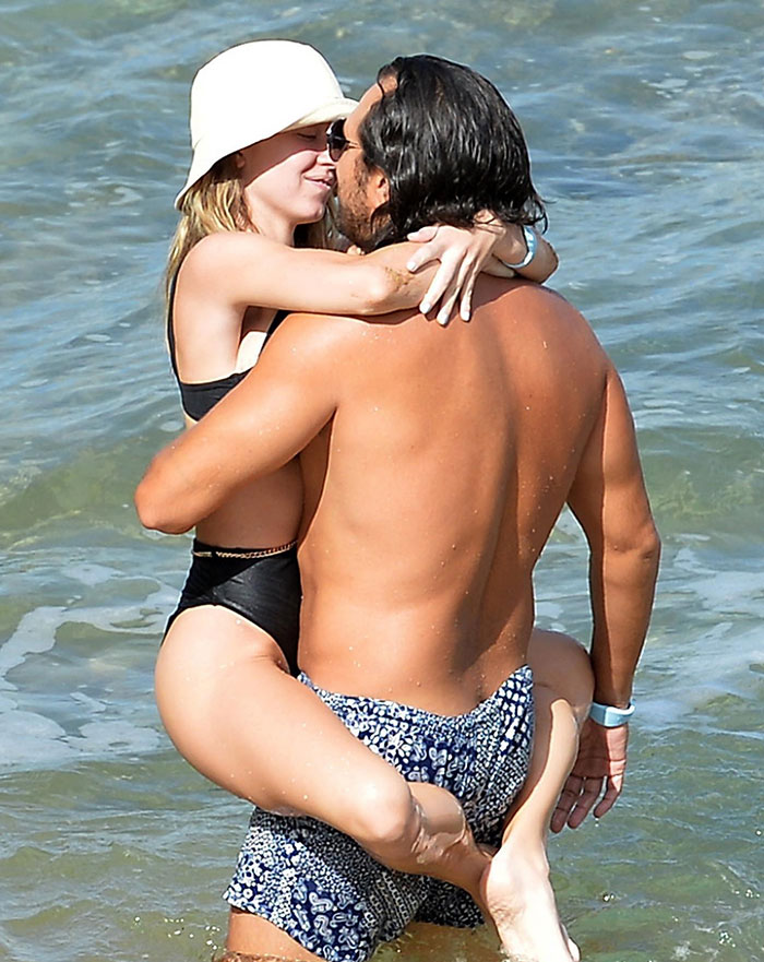 Sydney Sweeney, wearing a white sun hat and a black swimsuit, is held in the arms of Jonathan Davino, who is shirtless and wearing patterned swim trunks. They embrace affectionately in the water, smiling at each other. Sydney Sweeney, wearing a white sun hat and a black swimsuit, is held in the arms of Jonathan Davino, who is shirtless and wearing patterned swim trunks. They embrace affectionately in the water, smiling at each other.
