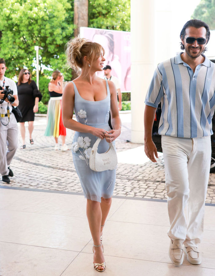 Sydney Sweeney, in a light blue floral dress with a white handbag, walks beside Jonathan Davino, who wears a blue striped shirt and white pants, at an outdoor event. Sydney Sweeney, in a light blue floral dress with a white handbag, walks beside Jonathan Davino, who wears a blue striped shirt and white pants, at an outdoor event.